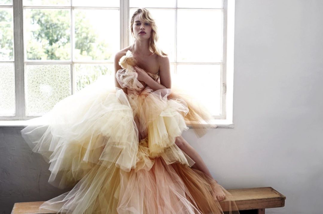 Young woman in a strapless dress made of layered tulle sitting on a wooden bench near a window in a bright, natural light setting.