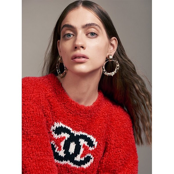 Close-up of a woman with brown hair and blue eyes wearing statement hoop earrings and a red sweater with a logo. Fashion shot.