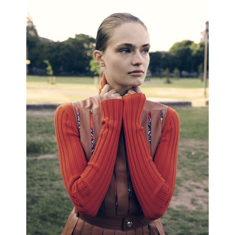 Woman in orange ribbed sweater poses in a park with grass and trees in the background. The photo captures a fashion editorial moment.