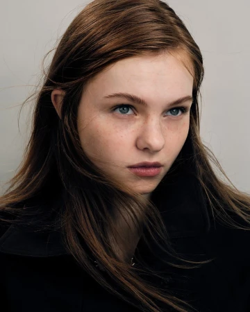 Close-up portrait of a young woman with long brown hair and blue eyes wearing a black jacket against a grey background.
