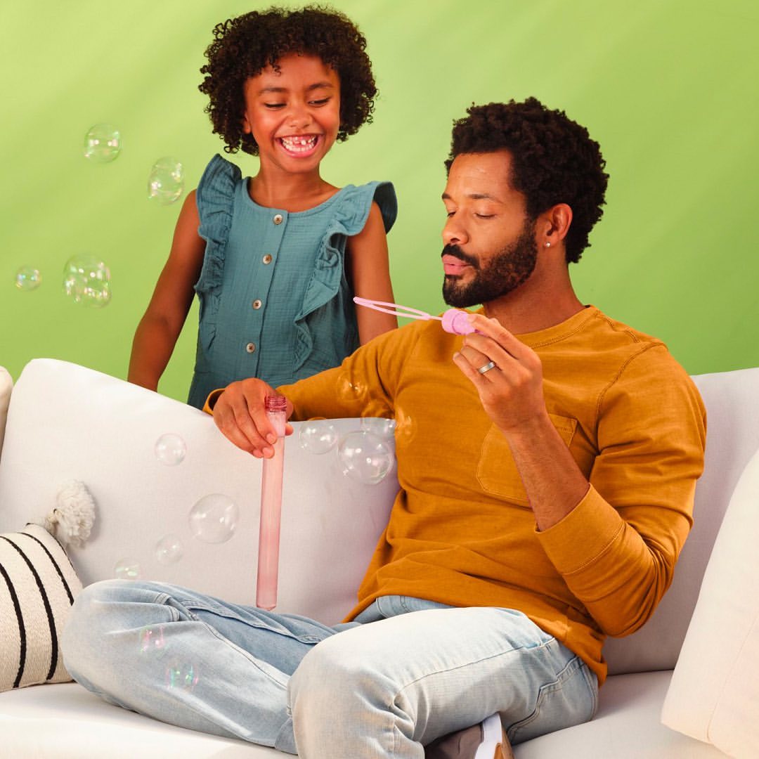 A happy African American father and daughter sit on a couch blowing bubbles indoors with a light green background, smiling and enjoying family time.