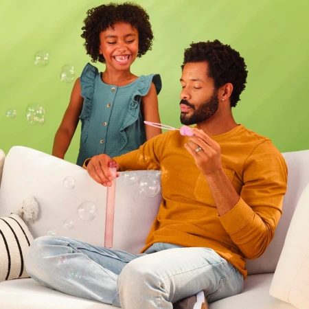 A happy African American father and daughter sit on a couch blowing bubbles indoors with a light green background, smiling and enjoying family time.