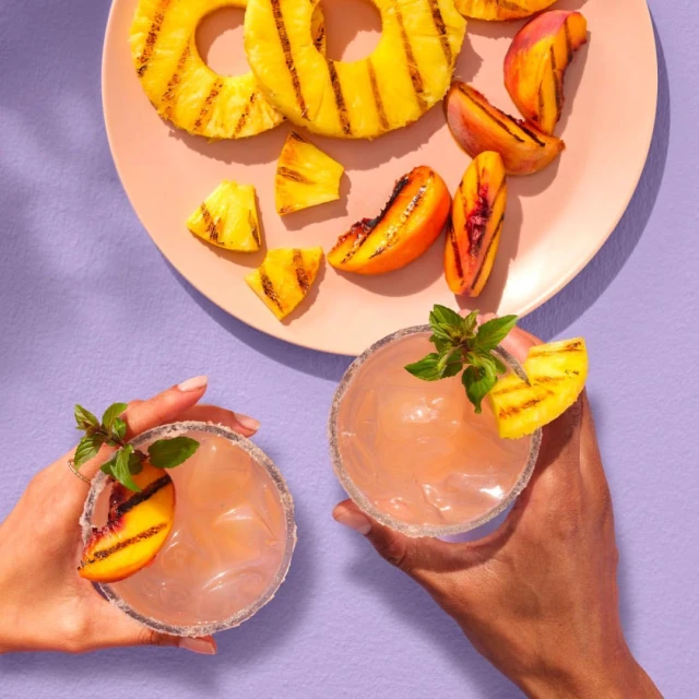 Overhead shot of two hands holding drinks with grilled peach and pineapple garnish, with grilled pineapple and peaches on a pink plate.