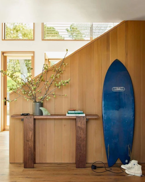 Interior view of a modern home with a blue surfboard, a wooden console table with decorations, and stairs with wood paneling.