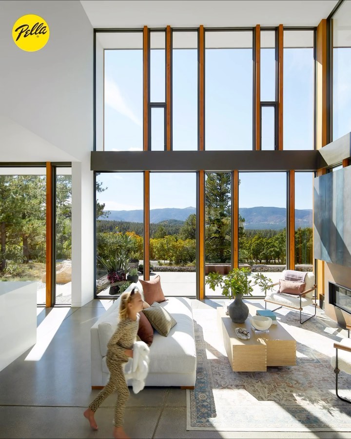 A young child runs through a modern living room with large windows revealing a scenic view of trees and hills on a sunny day.