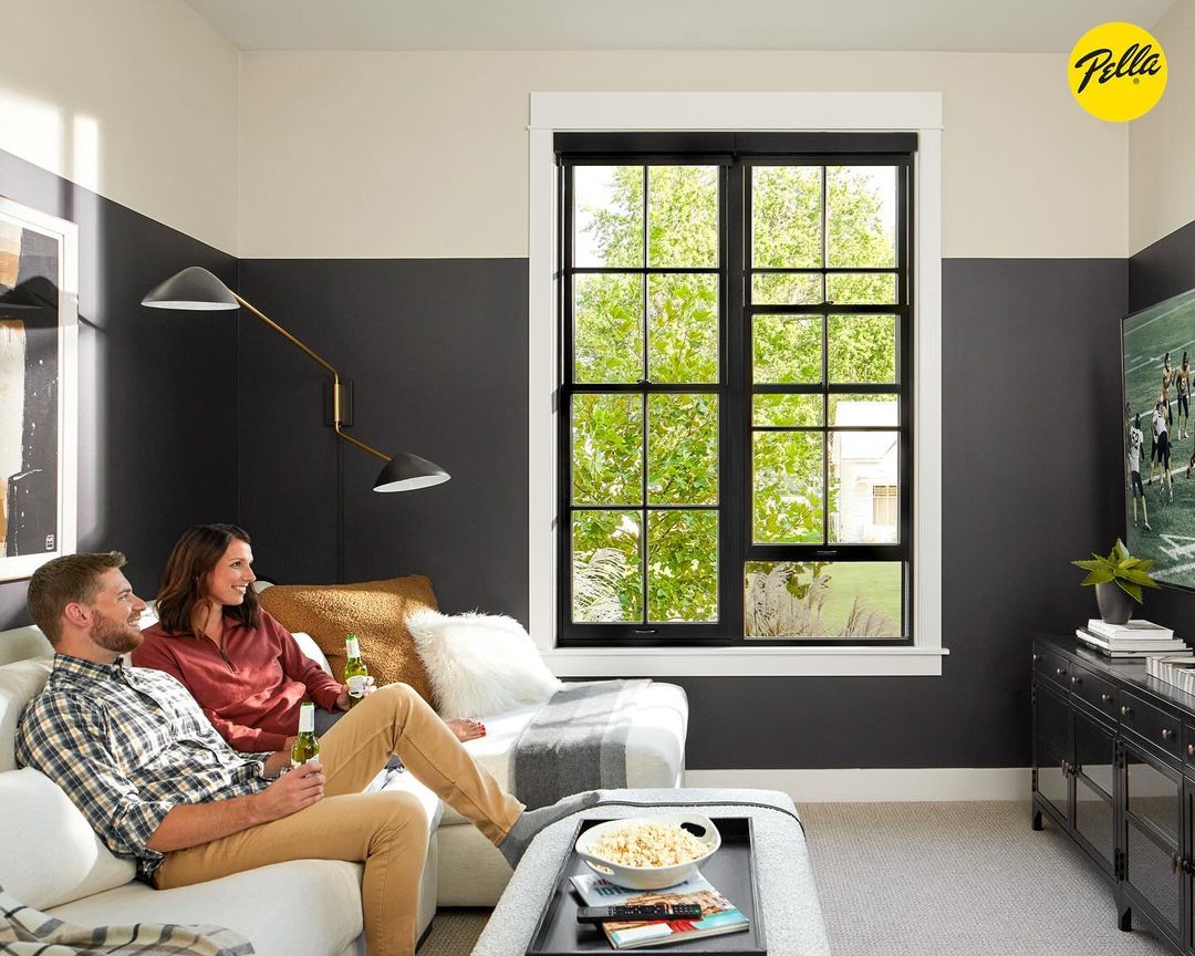 A couple relaxes on a white sofa in a contemporary living room with a large window, watching television and enjoying drinks.