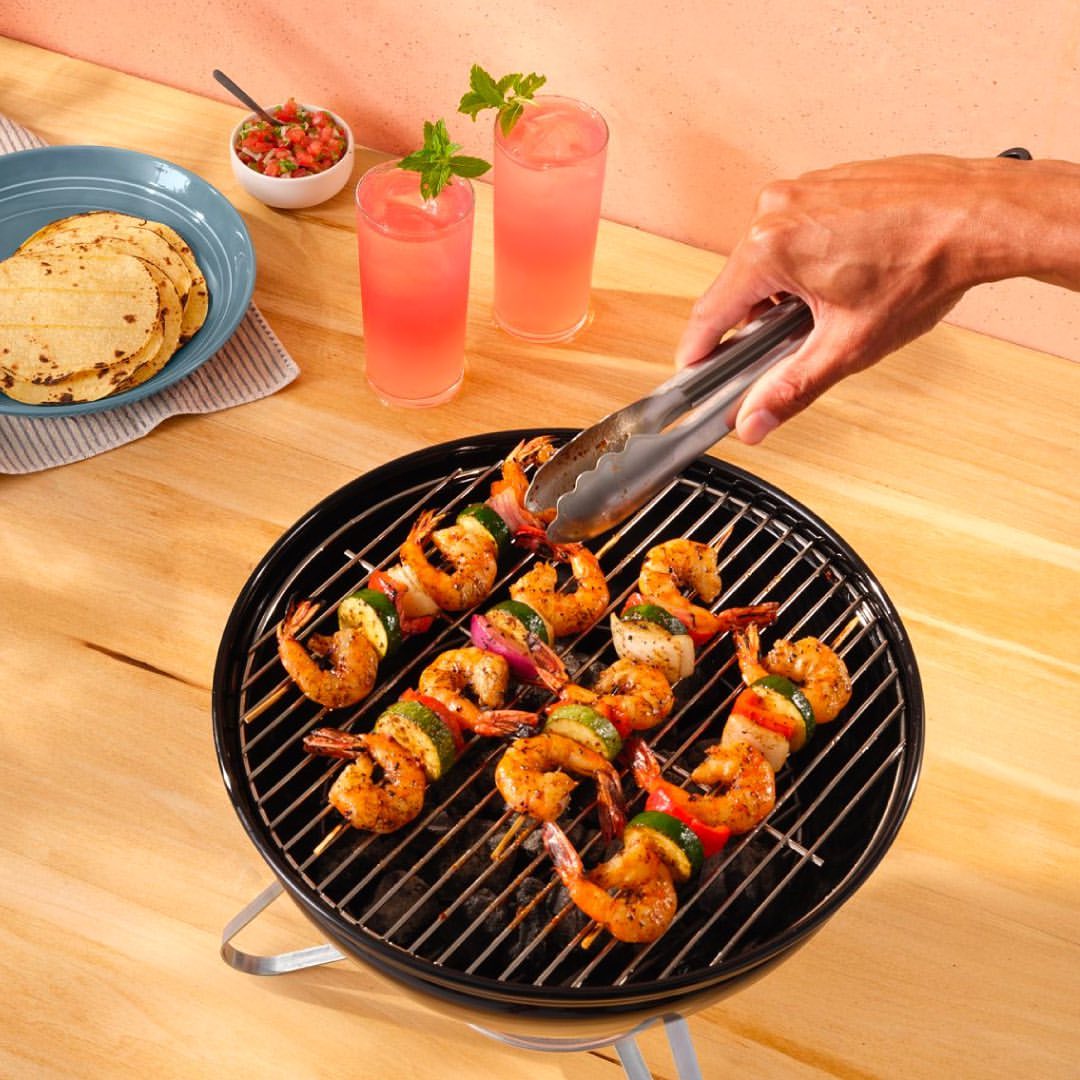 Grilled shrimp skewers with vegetables on a small grill, served with salsa, tortillas, and pink cocktails on a wooden table.