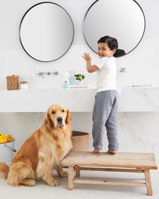 Young child stands on a wooden stool in a bright bathroom with a Golden Retriever dog nearby, next to a modern marble sink.