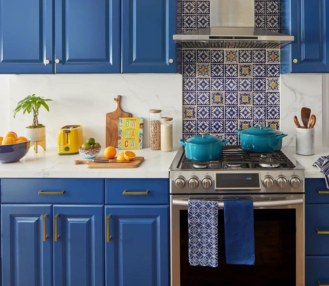Bright kitchen interior featuring blue cabinetry, stainless steel appliances, and a colorful tiled backsplash. Countertop displays fresh produce.