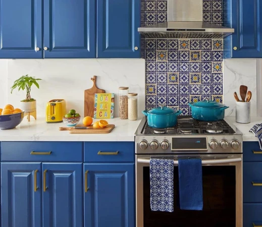 Bright kitchen interior featuring blue cabinetry, stainless steel appliances, and a colorful tiled backsplash. Countertop displays fresh produce.