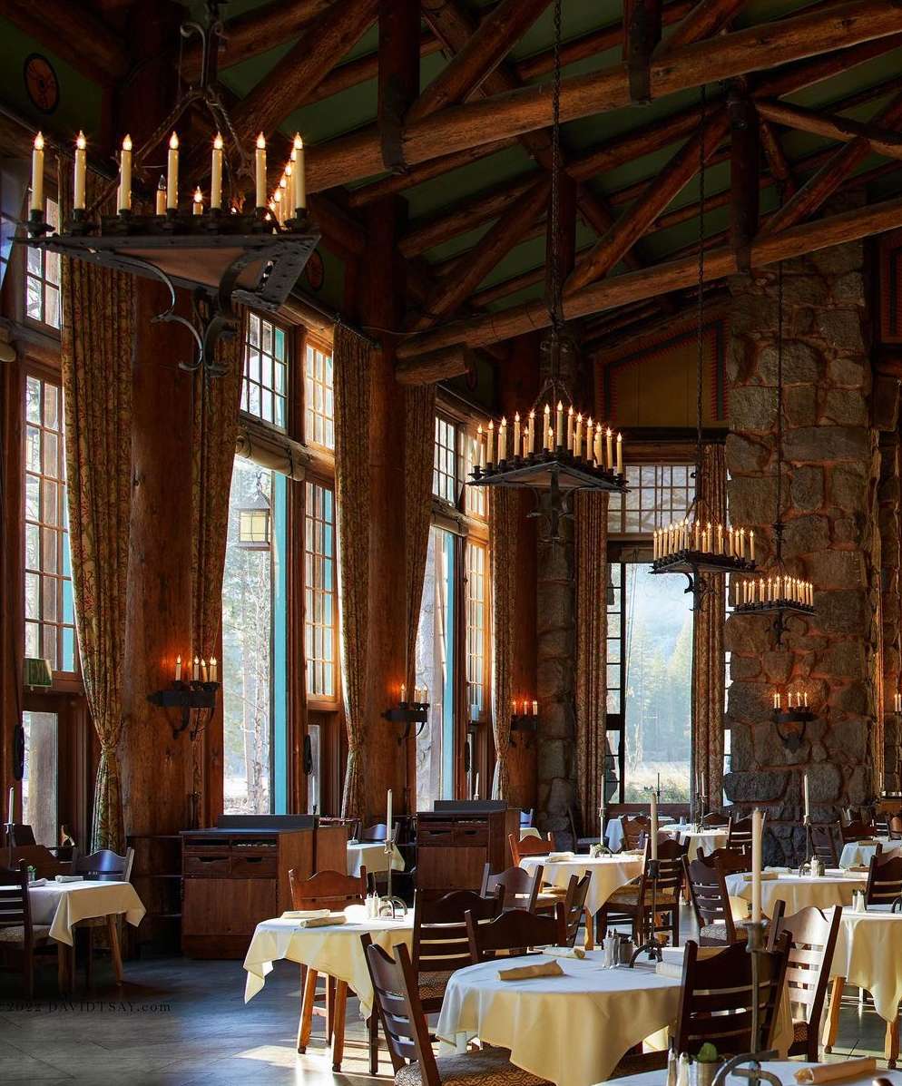 Interior of a grand dining hall with large chandeliers, set tables with white tablecloths, wooden chairs, and tall windows.