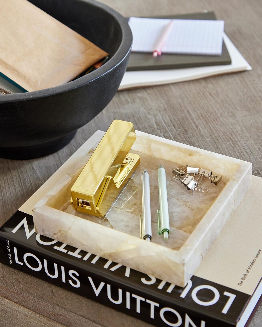Stationery set with a gold stapler, pens, and paperclips on a white marble tray, placed on a fashion book atop a wooden surface.