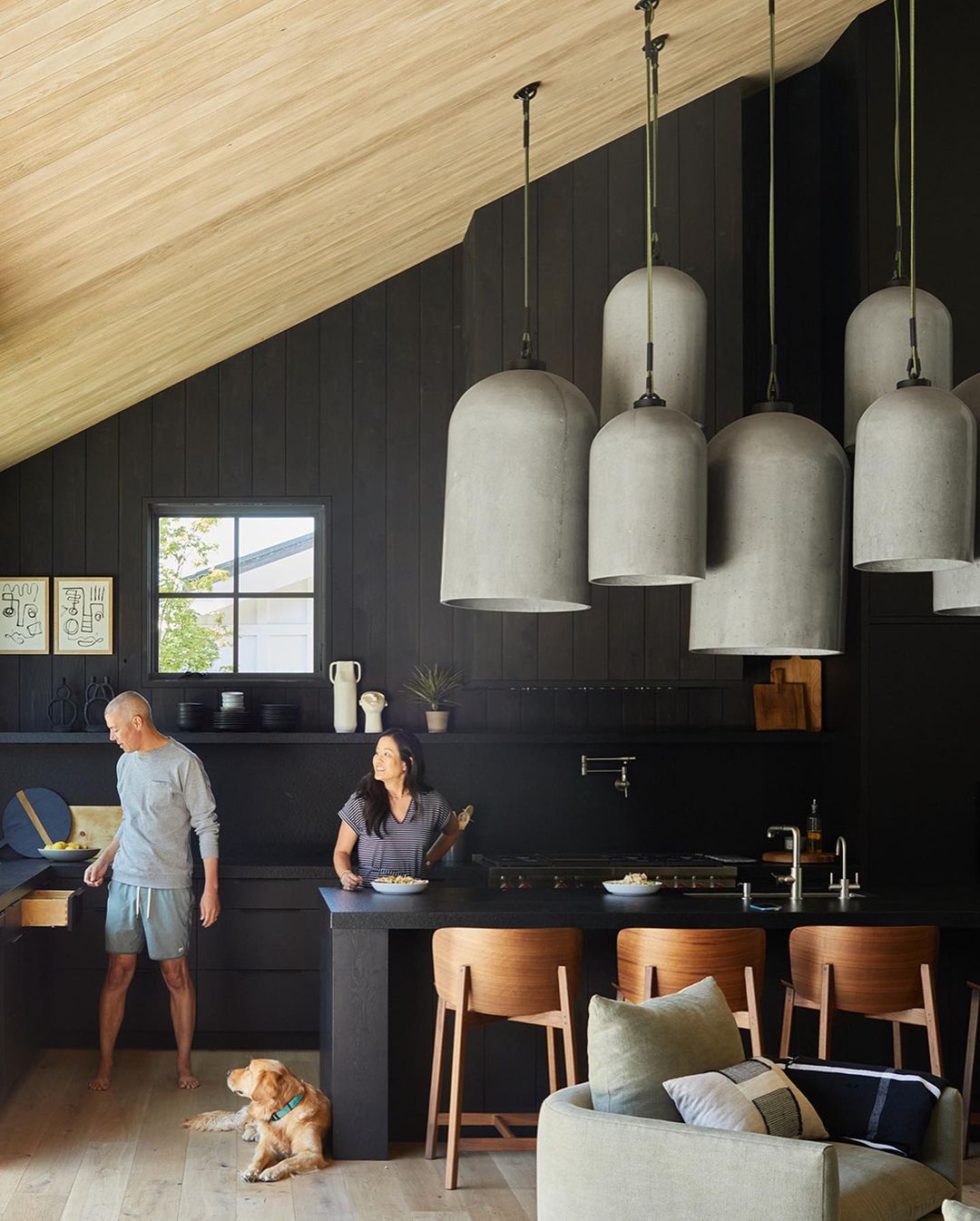 Contemporary black kitchen with two adults, a golden retriever, pendant lights, wood accents, and architectural elements.