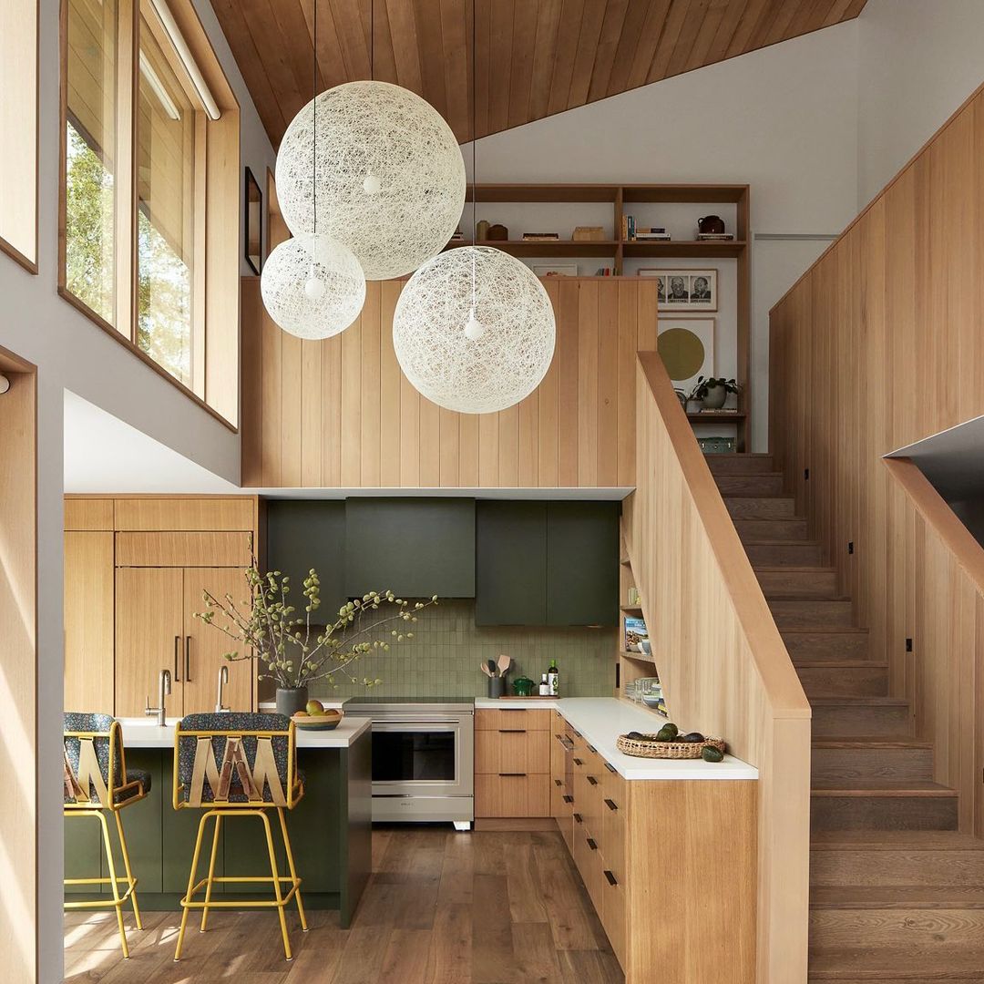 Contemporary interior with wooden walls, stylish bar stools, a kitchen with olive green cabinets, and decorative spherical lighting.