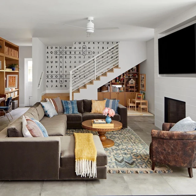 Living room interior with a brown sectional sofa, patterned rug, coffee table, TV, and a decorative word-search wall design.