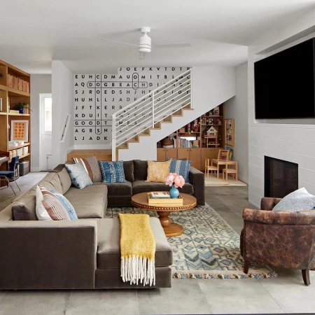 Living room interior with a brown sectional sofa, patterned rug, coffee table, TV, and a decorative word-search wall design.