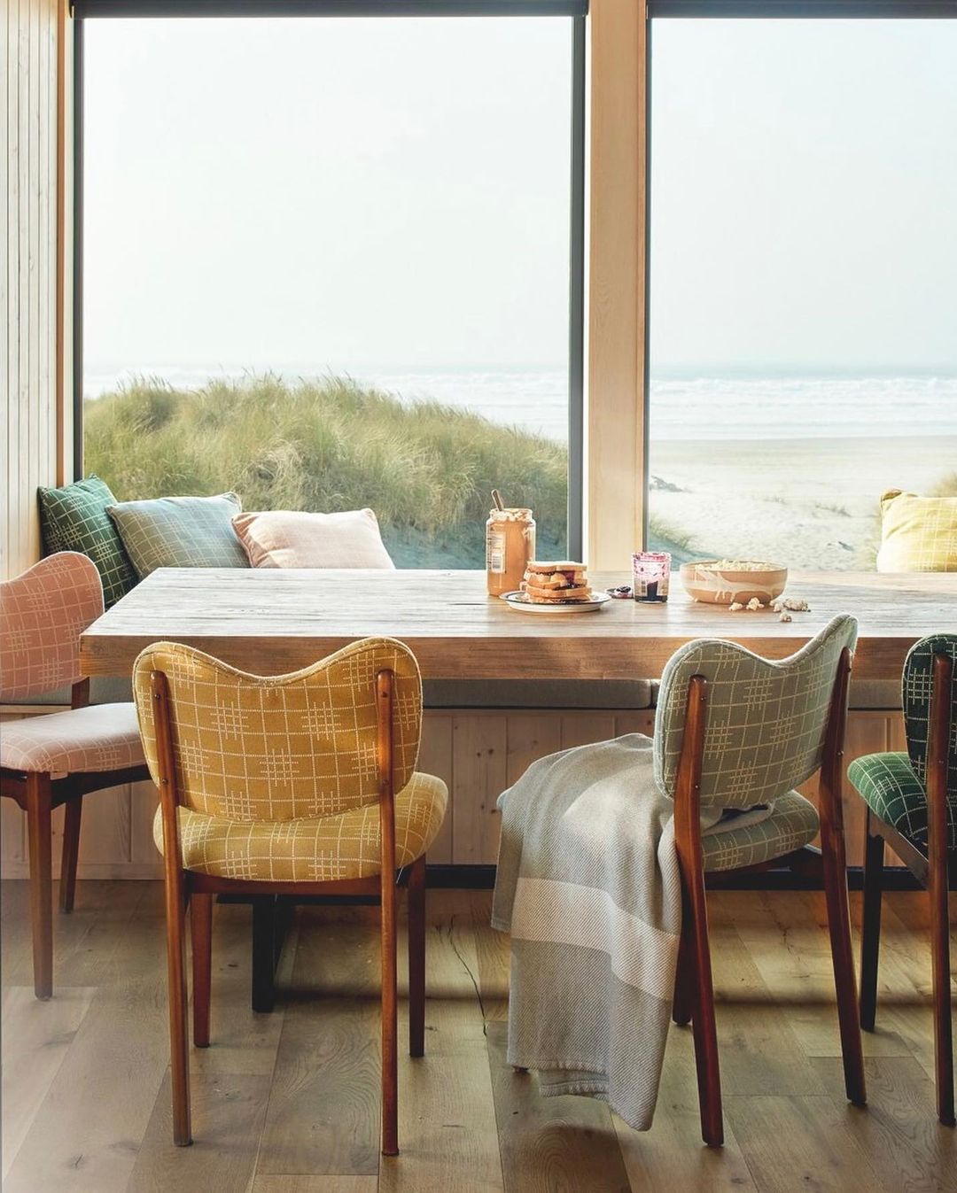 Dining room interior with wooden table, patterned chairs, and coastal view through large window, evoking a cozy and stylish atmosphere.