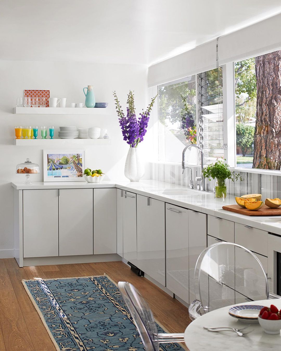 Bright, modern kitchen interior with white cabinets, marble countertops, blue rug, large windows, and vase of purple flowers.