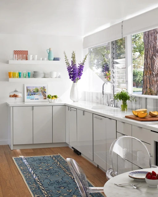 Bright, modern kitchen interior with white cabinets, marble countertops, blue rug, large windows, and vase of purple flowers.