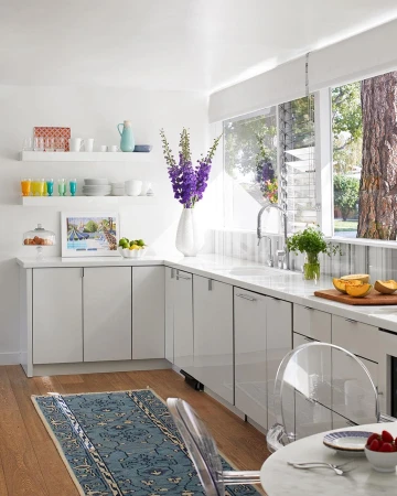Bright, modern kitchen interior with white cabinets, marble countertops, blue rug, large windows, and vase of purple flowers.