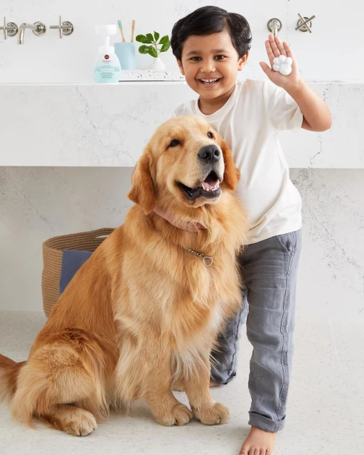 Child and golden retriever dog pose in a bright bathroom, holding up a hand covered in white soap bubbles for cleaning and hygiene.