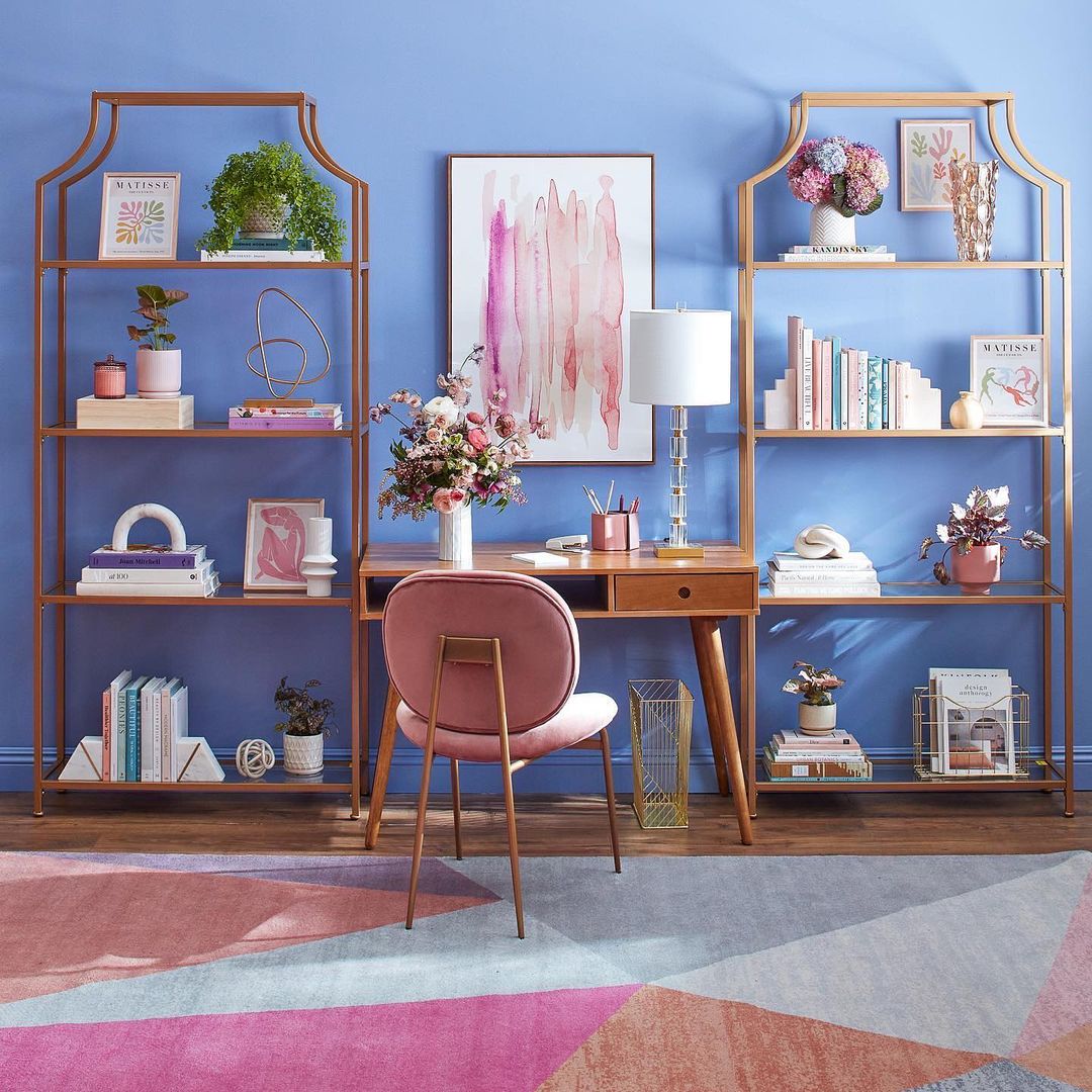 Home office interior featuring a desk, pink chair, open shelving units displaying decor, and a geometric-patterned area rug.