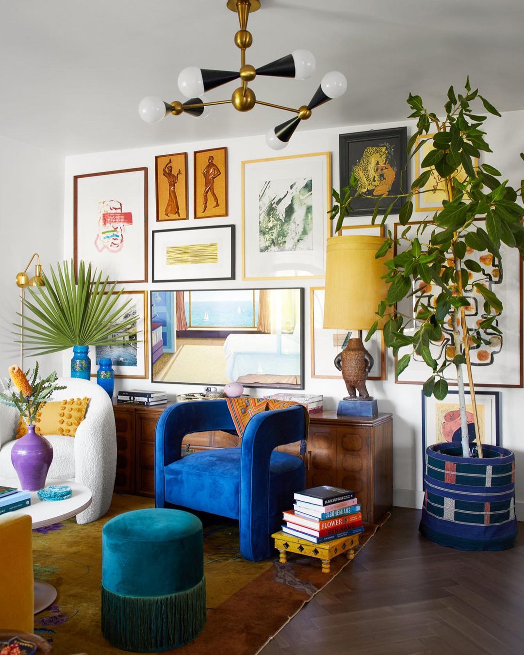 Eclectic living room interior with blue velvet chair, ottoman, sofa, and a gallery wall with colorful artwork and modern chandelier.