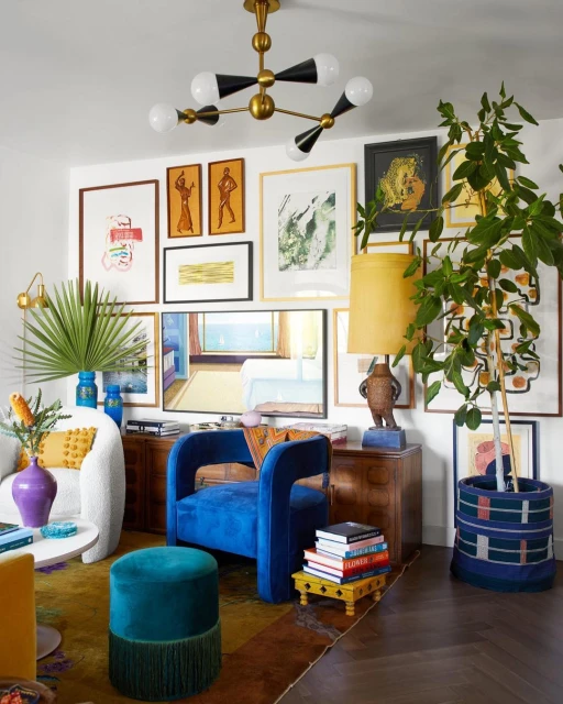 Eclectic living room interior with blue velvet chair, ottoman, sofa, and a gallery wall with colorful artwork and modern chandelier.