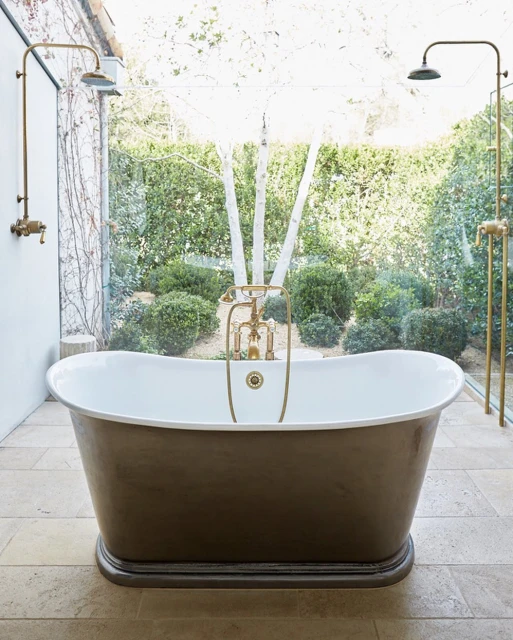 Freestanding bathtub with brass fixtures inside bright bathroom, garden view. Interior design, home decor, luxury bathroom.