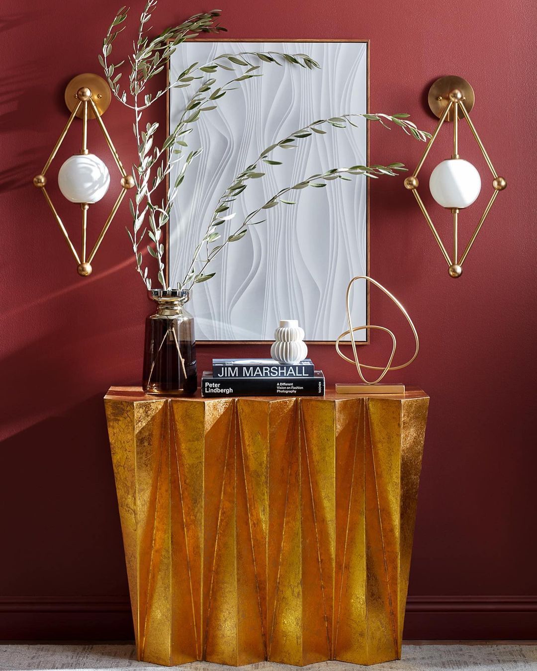 Interior space features a golden console table with a vase, books, abstract sculpture, and two sconces against a dark red wall.