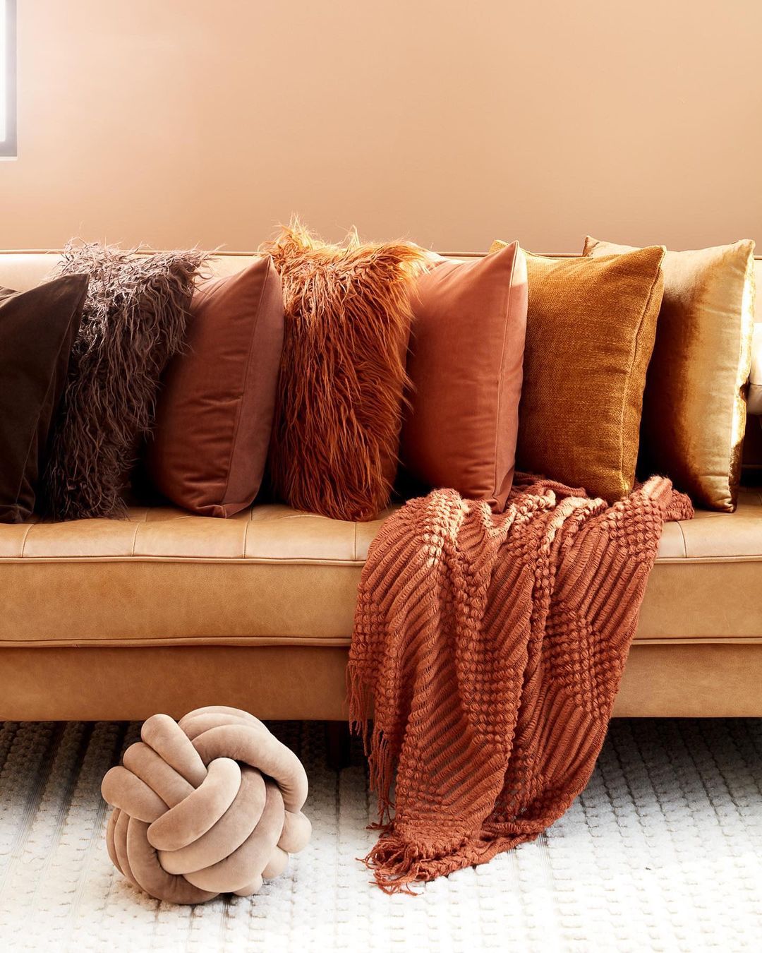 Still life of decorative pillows on a caramel-colored couch against a peach-toned wall, featuring a knitted throw and knot pillow.