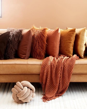 Still life of decorative pillows on a caramel-colored couch against a peach-toned wall, featuring a knitted throw and knot pillow.