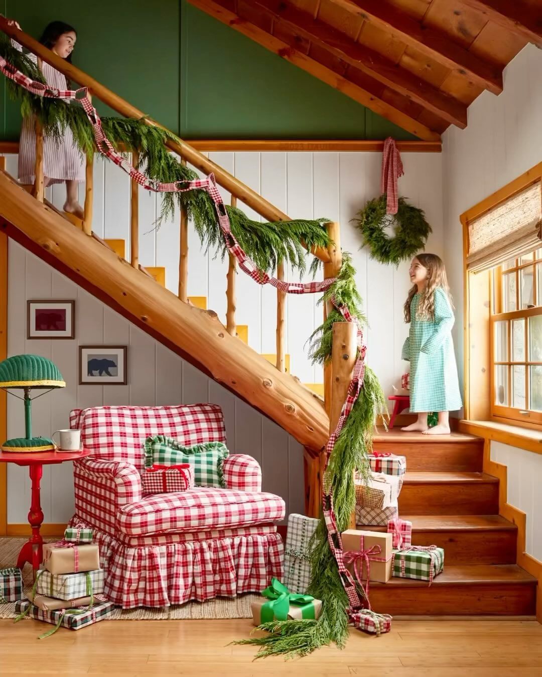 Two young girls stand near a staircase in a cozy home interior decorated with holiday decor, presents, and an armchair with a red plaid pattern.