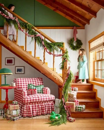 Two young girls stand near a staircase in a cozy home interior decorated with holiday decor, presents, and an armchair with a red plaid pattern.