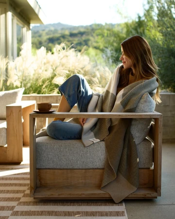 Woman wrapped in a patterned blanket seated in an outdoor wooden armchair holding a ceramic cup