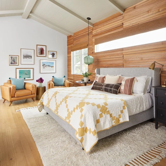 Bedroom interior with a light gray bed covered in a patterned quilt, accented by throw pillows, with wood paneled wall and art gallery.