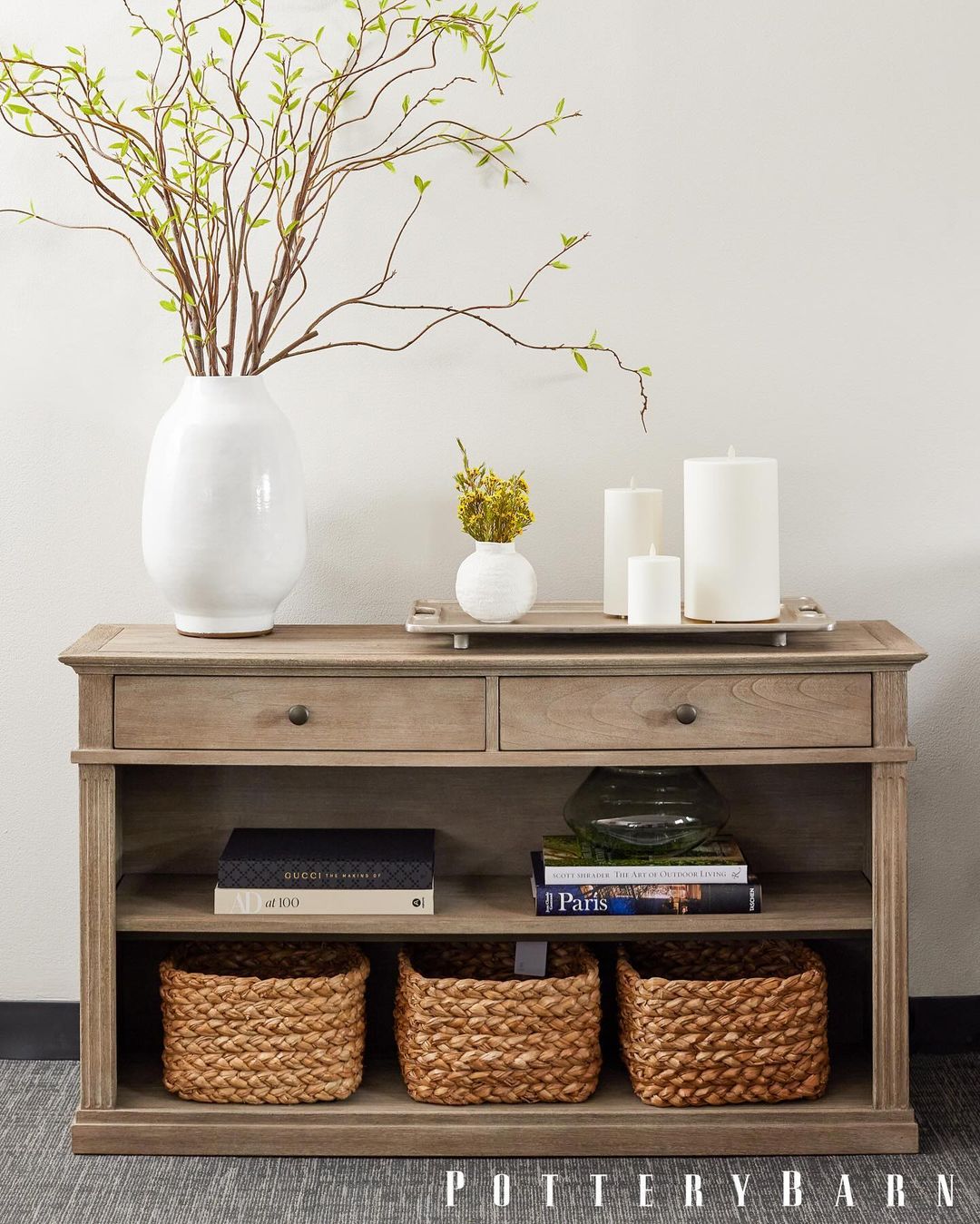 Interior view of a console table with decorative accents: vases with branches, candles, books, and woven baskets beneath.