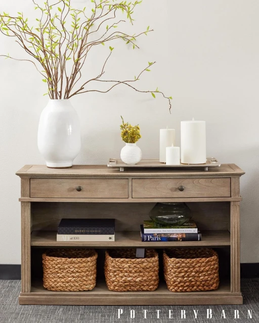 Interior view of a console table with decorative accents: vases with branches, candles, books, and woven baskets beneath.