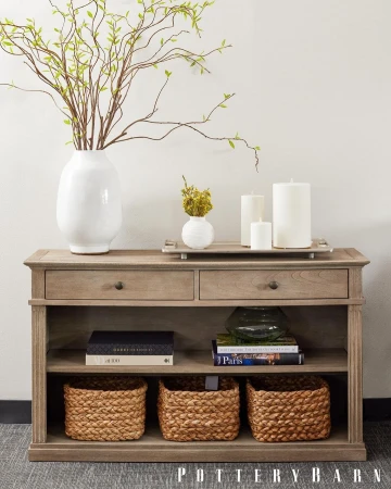 Interior view of a console table with decorative accents: vases with branches, candles, books, and woven baskets beneath.