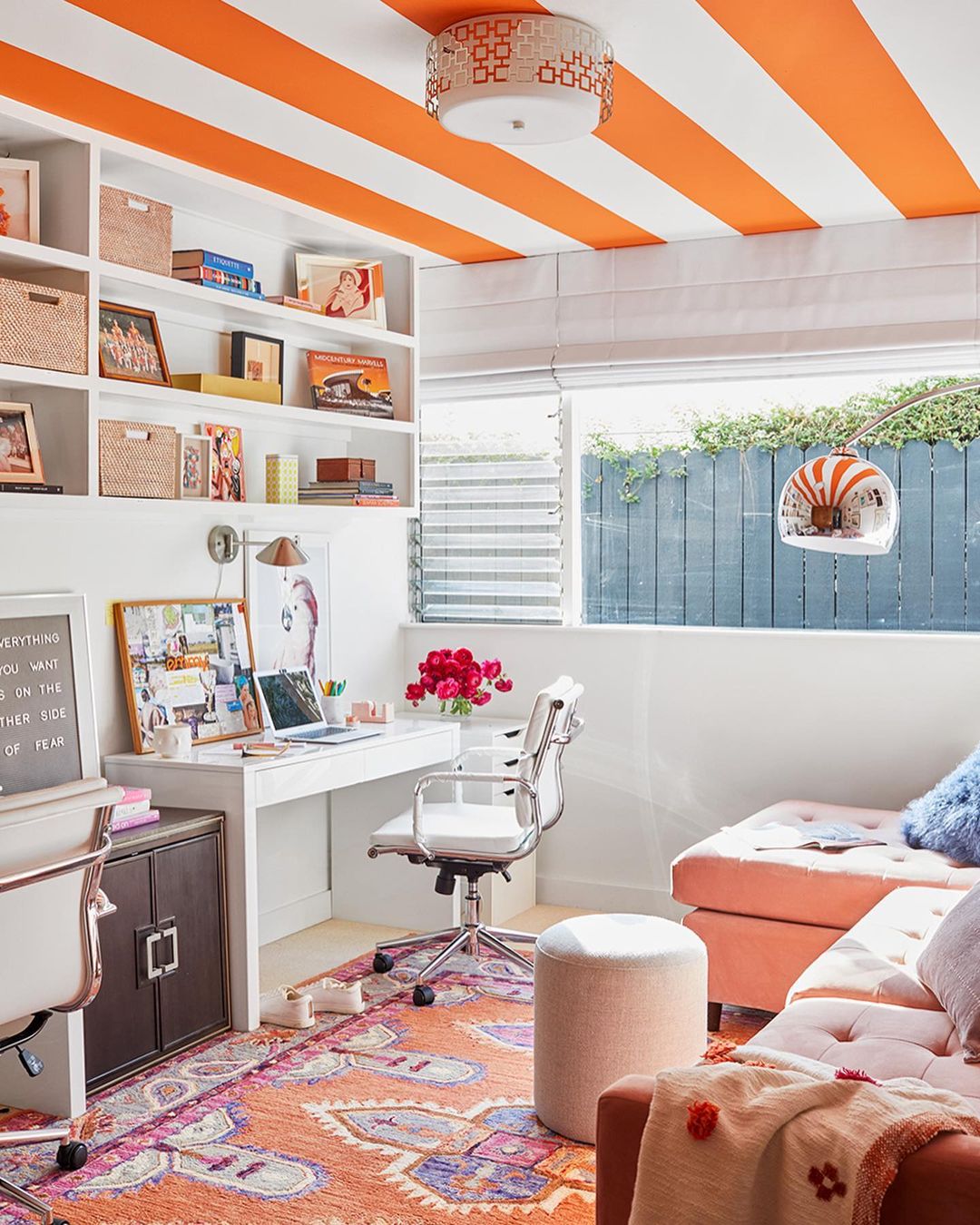 Brightly decorated home office interior with white desk, shelves, and accent seating, featuring orange and white striped ceiling and colorful rug.