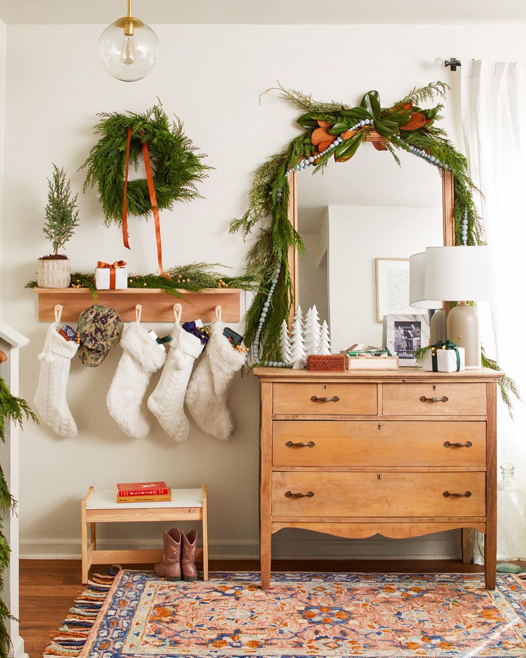 Festive holiday scene with a wooden dresser adorned with Christmas greenery, stockings hung on a wall shelf, and patterned rug.