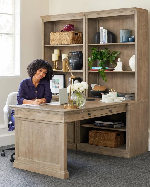 Woman works at a light wood desk with a hutch holding decor; a laptop, lamp and flowers sit on the desk in an indoor setting.