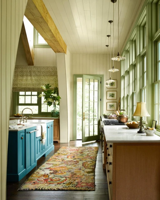 Bright, naturally lit eat-in kitchen with a blue kitchen island, colorful Persian rug, white marble countertops and green framed windows.