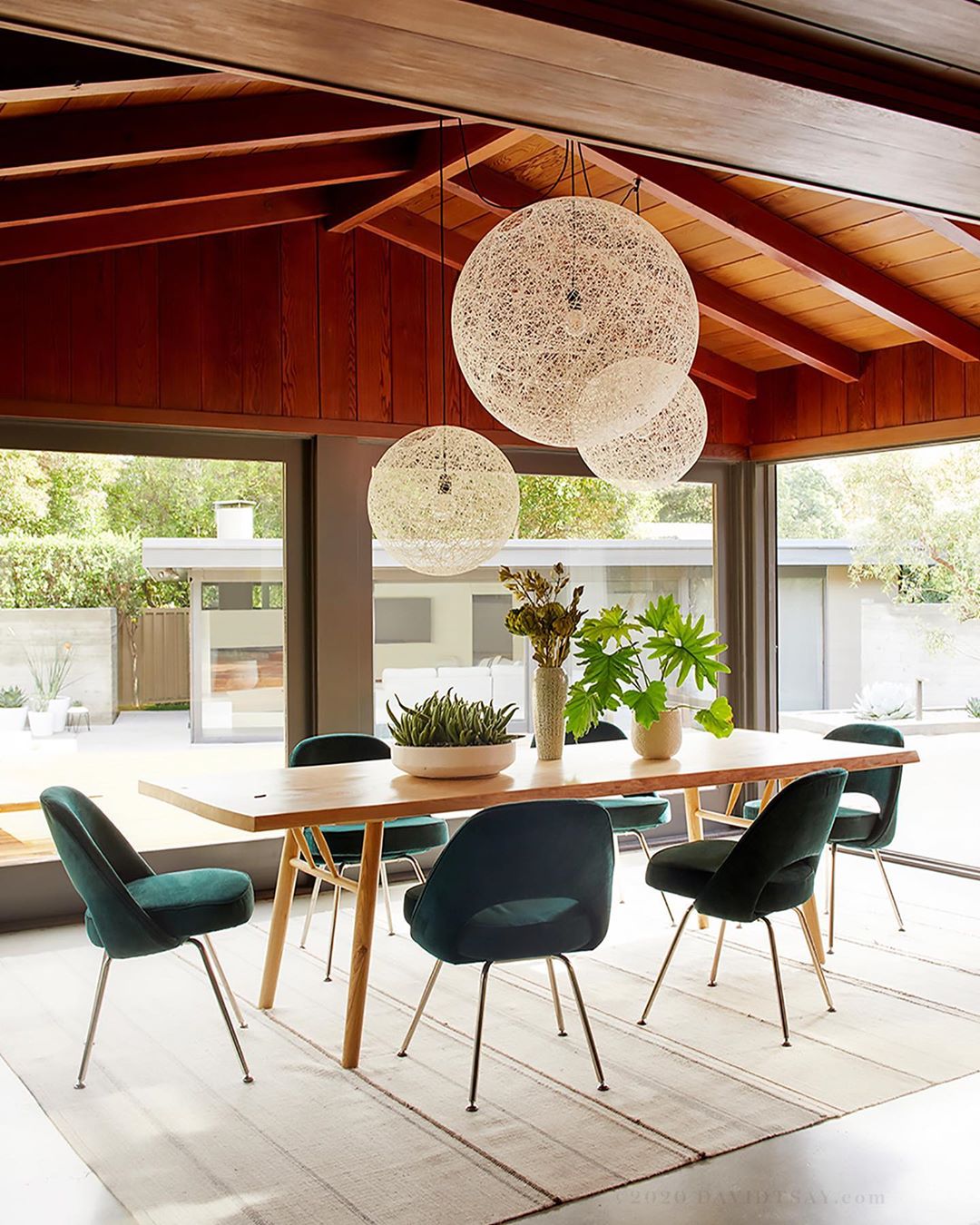 Modern dining room interior with wooden table, green velvet chairs, and spherical pendant lights, bathed in natural daylight.