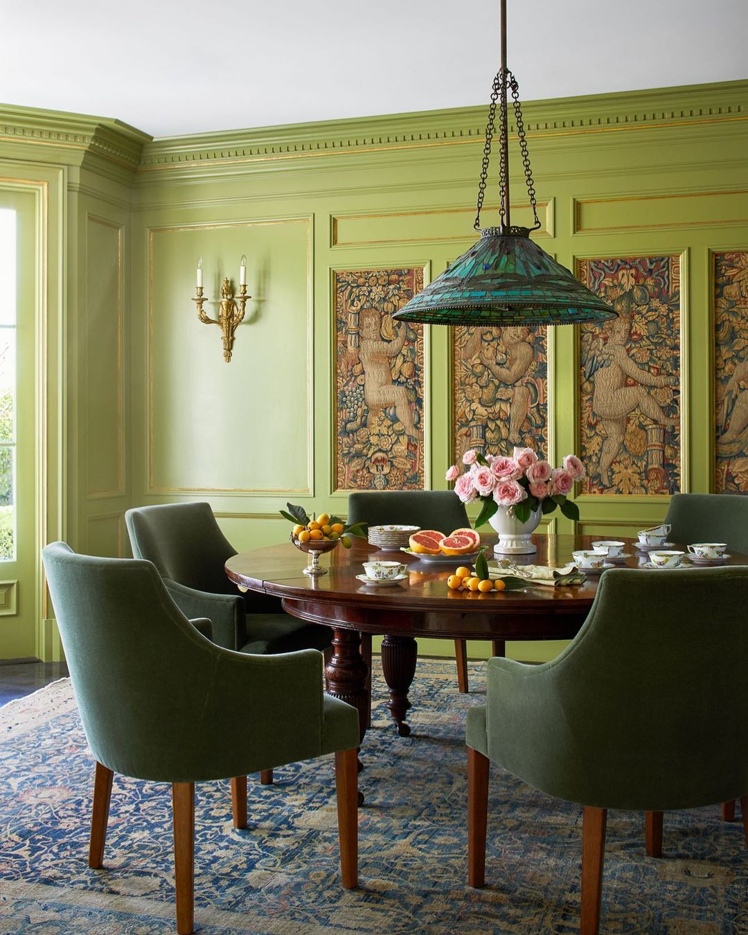 Elegant dining room featuring a dark wood table set with china and fruit, surrounded by green upholstered chairs on a patterned rug.