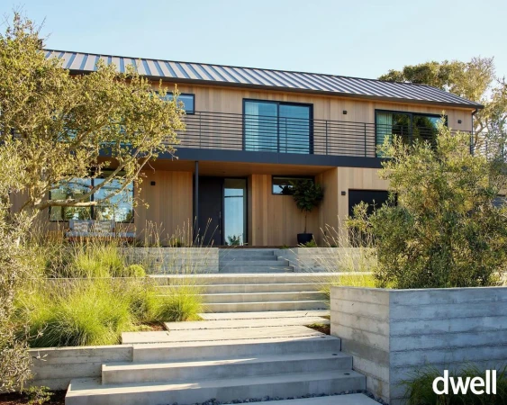 Exterior of modern two-story home with wood siding, metal roof, concrete stairs, and green landscaping on a sunny day.