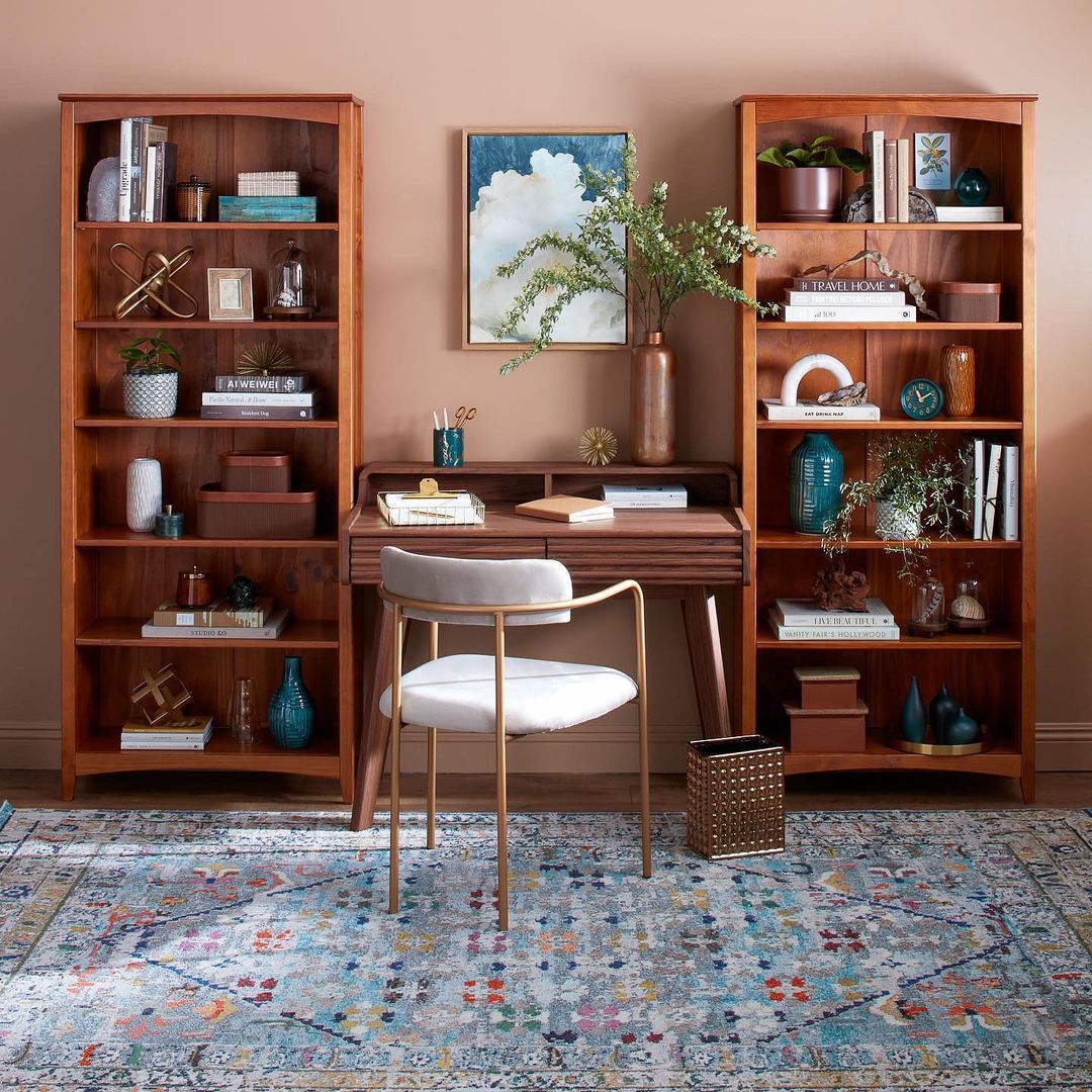 Home office setup with wooden desk and chair, bookshelves with decor, and patterned rug, creating a cozy interior design.