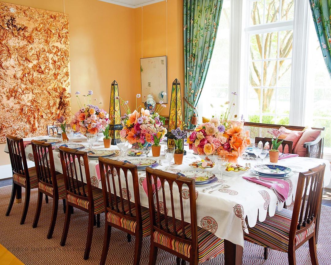 Dining room interior featuring a long table set with floral centerpieces, plates, and linens, surrounded by wooden chairs and decor.