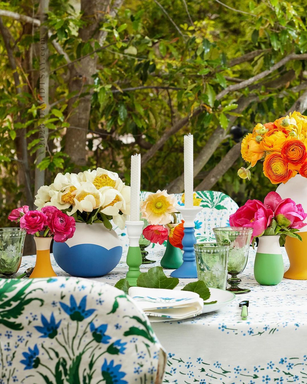 Outdoor tablescape with a white floral patterned tablecloth, colorful floral arrangements, and candlesticks in bright blue, yellow, and green.