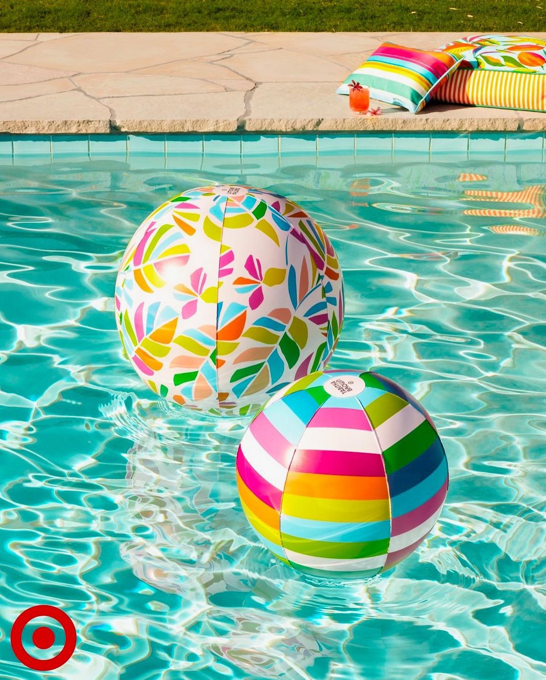 Two inflatable beach balls float in a bright blue swimming pool next to colorful striped pillows on a stone patio with grass.